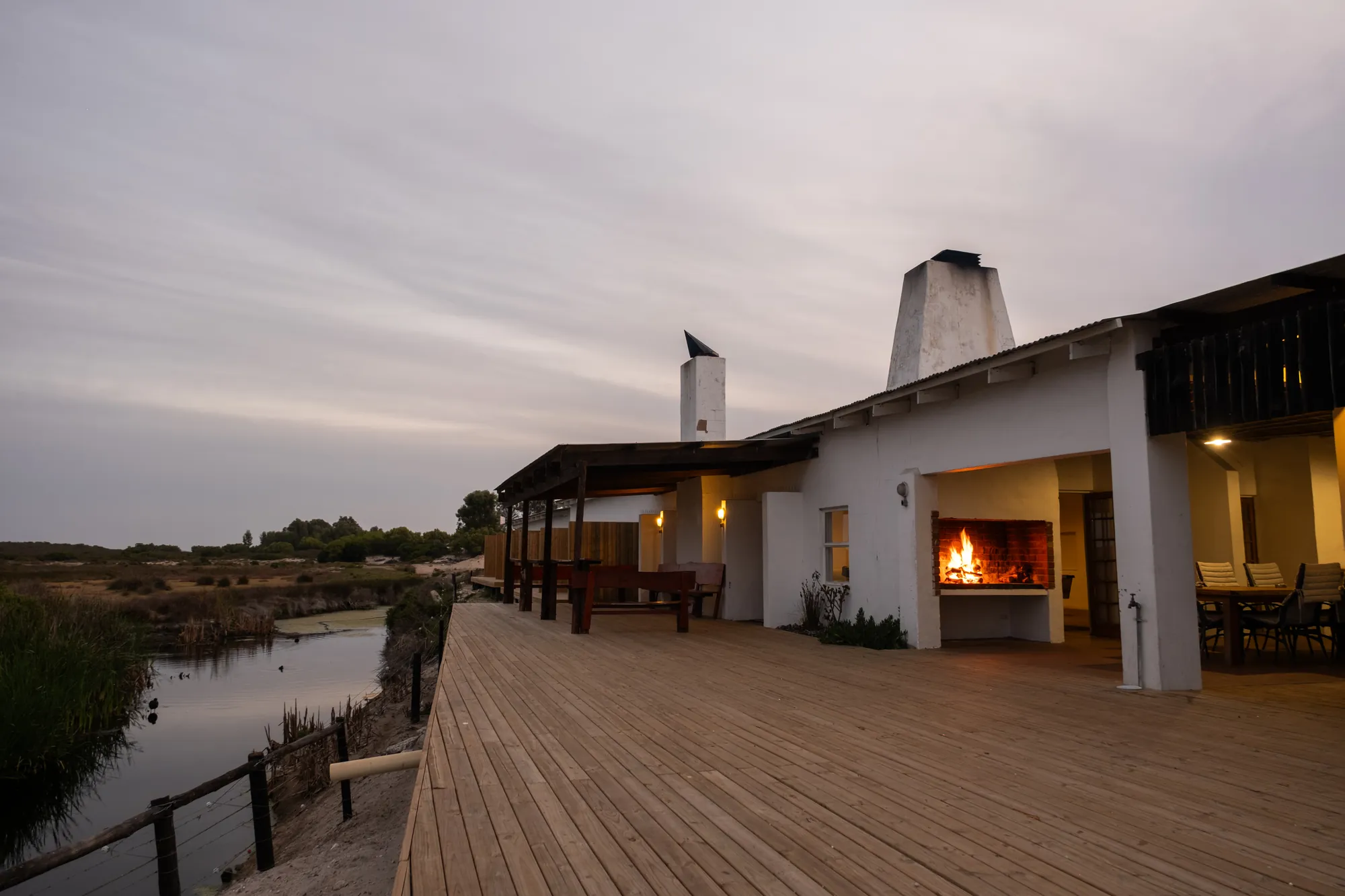 The long veranda of Hide-Away Lodge with a fire burning, beside a dam at dusk